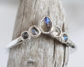 Puede incluir: Un anillo de plata con diseño de corona, con cinco piedras preciosas iridiscentes. Las piedras muestran tonos de azul y gris, engastadas en una banda de plata texturizada. El anillo se exhibe sobre un trozo de madera envejecida.