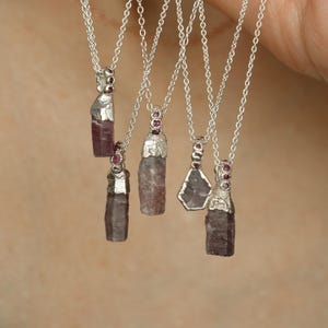 May include: A collection of silver necklaces with raw, natural gemstone pendants. Each pendant is a unique shape and color, ranging from light to dark purple, with silver accents and small red gemstones. The necklaces hang from delicate silver chains.