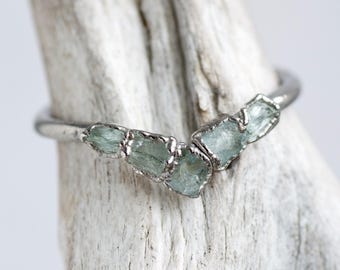 Puede incluir: Un anillo de plata con cinco piedras de aguamarina azul claro. Las piedras están engarzadas en una banda de plata, creando un aspecto único y natural. El anillo se muestra sobre un trozo de madera clara.