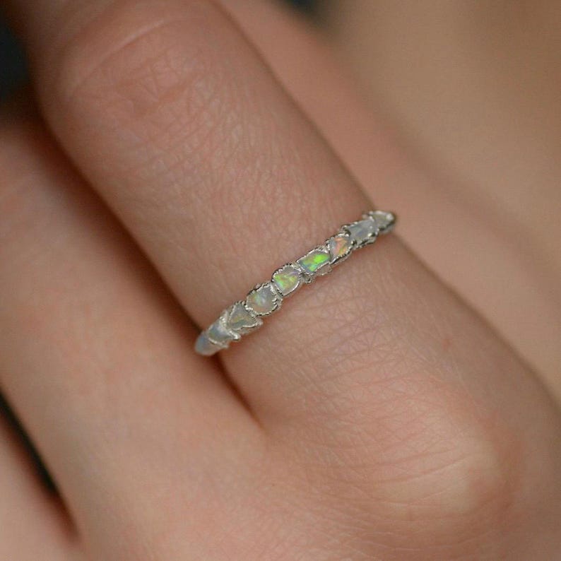 Op de afbeelding: Een zilveren ring met een rij kleine, ovale opalen. De opalen hebben een melkachtige witte basis met groene en gele flitsen. De ring wordt op een vinger getoond.