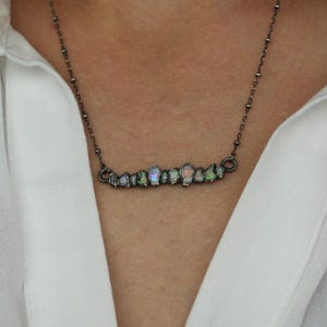 May include: A silver-toned necklace with a row of iridescent opal stones. The opals are set in a dark metal, creating a striking contrast. The necklace is worn against a white shirt, highlighting the jewelry's design.