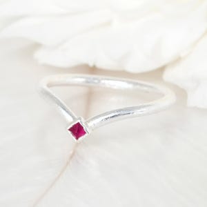 Può includere: Un anello d'argento con un design a forma di V. L'anello presenta una piccola gemma quadrata color rubino al centro. L'anello è posto su uno sfondo bianco morbido.