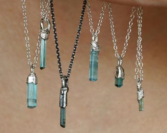 May include: Several silver necklaces with light blue crystal pendants. The pendants are cylindrical and vary in size. The chains have different styles, some with a darker, oxidised finish. The necklaces are displayed together.