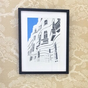 May include: A black and white drawing of a white building with a blue sky in the background. The building has many windows and a balcony. The drawing is framed in a black frame.