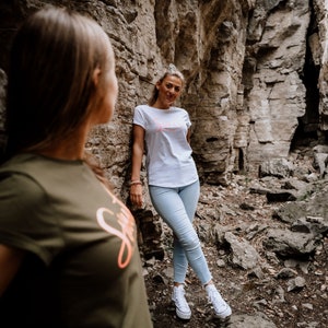 Może przedstawiać: Dwie osoby stojące w skalistym, zewnętrznym otoczeniu. Osoba po lewej stronie nosi ciemnozielony t-shirt z r&oacute;żowym nadrukiem. Osoba po prawej stronie nosi biały t-shirt z r&oacute;żowym nadrukiem i jasnoniebieskie dżinsy.