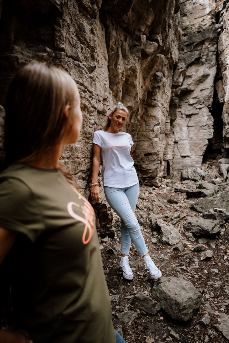Może przedstawiać: Dwie osoby stojące w skalistym, zewnętrznym otoczeniu. Osoba po lewej stronie nosi ciemnozielony t-shirt z r&oacute;żowym nadrukiem. Osoba po prawej stronie nosi biały t-shirt z r&oacute;żowym nadrukiem i jasnoniebieskie dżinsy.