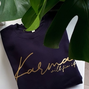 Op de afbeelding: Een marineblauwe sweatshirt met de gouden tekst "Karma will fix it". De crew neck sweatshirt is maat M en wordt gedeeltelijk omlijst door groene bladeren.