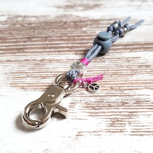 May include: A silver key chain with a silver clip, a gray and pink rope, a silver peace sign charm, and a gray bead with a paw print.