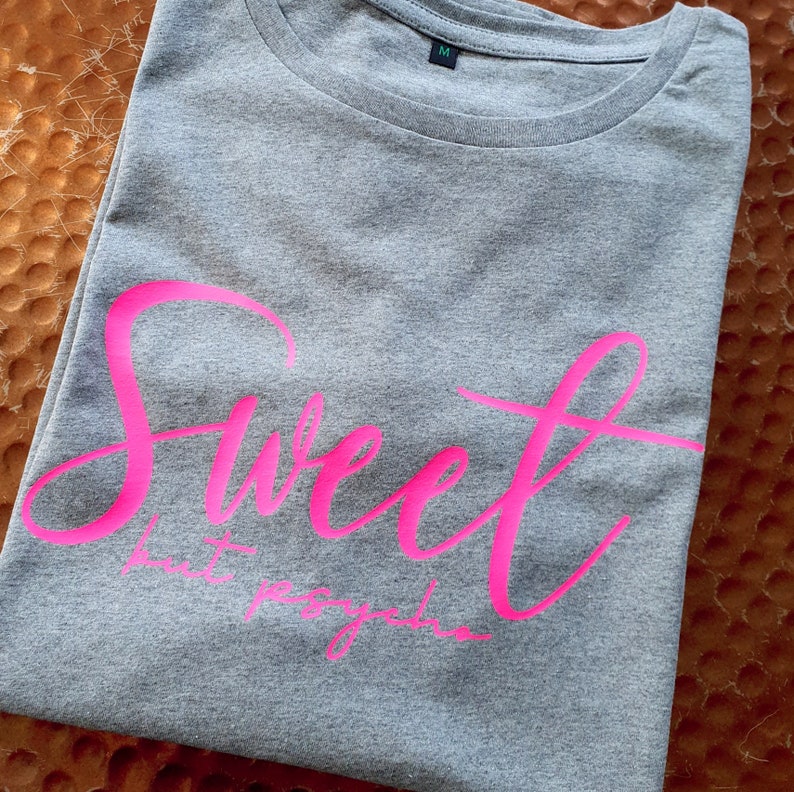Może przedstawiać: Szara koszulka z napisem "Sweet but psycho" w r&oacute;żowych literach.