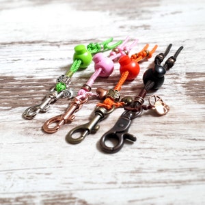 May include: Five colorful keychains with different charms. Each keychain has a metal clasp and a colorful bead. The charms include a paw print, a heart, a peace sign, and a skull.
