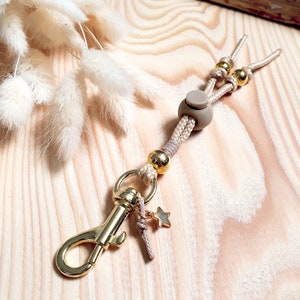 May include: A beige and gold keychain with a gold clasp, a beige rope, and gold beads. The keychain has a small star charm.