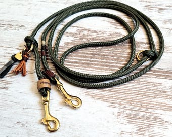 May include: A green rope dog leash with two gold metal clips and a brown leather accent. The leash has a small decorative charm with a heart and tassels.