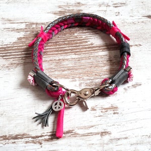 Può includere: Un collare per cani intrecciato rosa e grigio con un ciondolo a forma di segno della pace argentato e un fiocco grigio.