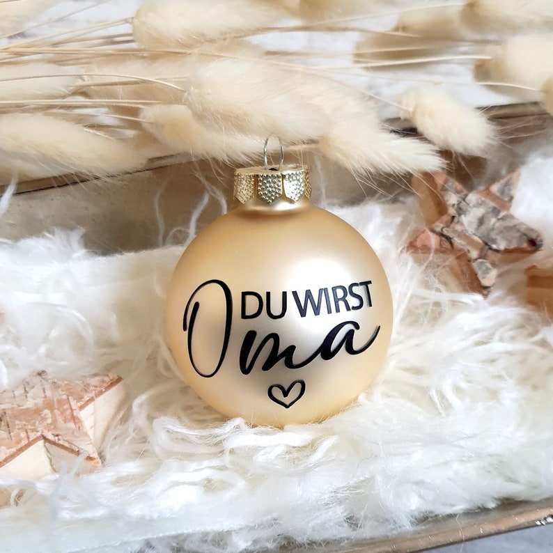May include: Gold Christmas ornament with the text "DU WIRST OMA" in black lettering and a small heart.