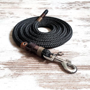 Op de afbeelding: Zwarte hondenriem met leren accenten en een zilverkleurige metalen sluiting. De riem is opgerold op een licht houten oppervlak. De riem heeft een gevlochten textuur en is voor honden.