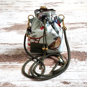 Könnte beinhalten: Eine kleine, kordelzugbeuteltasche mit einem grün-weißen Blumendruck mit Vögeln. Die Tasche hat einen grünen Seilriemen und goldfarbene Beschläge. Die Tasche hat ein Etikett mit der Aufschrift "felius".