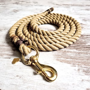 Op de afbeelding: Een bruin touw hondenriem met een goudkleurige sluiting en een kleine hartvormige bedel. De riem is opgerold op een lichtgekleurd houten oppervlak. Het touw is dik en lijkt duurzaam, geschikt voor hondenbezitters.