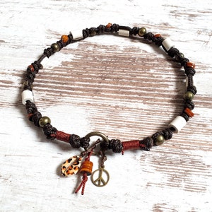 Puede incluir: Un collar de cuentas marrones, rojas y blancas con un dije de signo de paz y un dije de concha. El collar está hecho de cordón y tiene una variedad de cuentas, incluyendo cuentas pequeñas redondas, cuentas cilíndricas más grandes y algunas cuentas más grandes y planas.