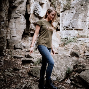 Może przedstawiać: Kobieta w zielonej koszulce z napisem "Sweet Escape" w pomarańczowych literach, niebieskich dżinsach i czarnych butach. Stoi na skalistym podłożu.