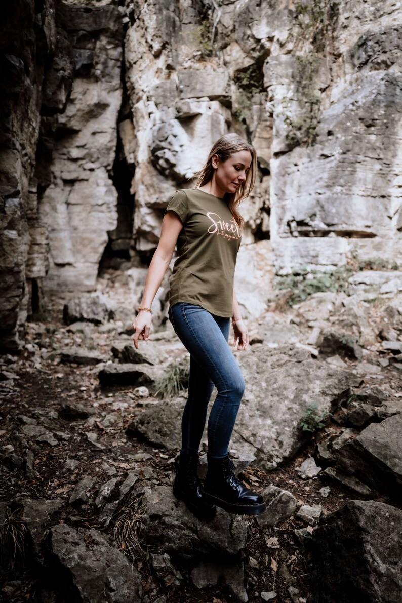 Może przedstawiać: Kobieta w zielonej koszulce z napisem "Sweet Escape" w pomarańczowych literach, niebieskich dżinsach i czarnych butach. Stoi na skalistym podłożu.