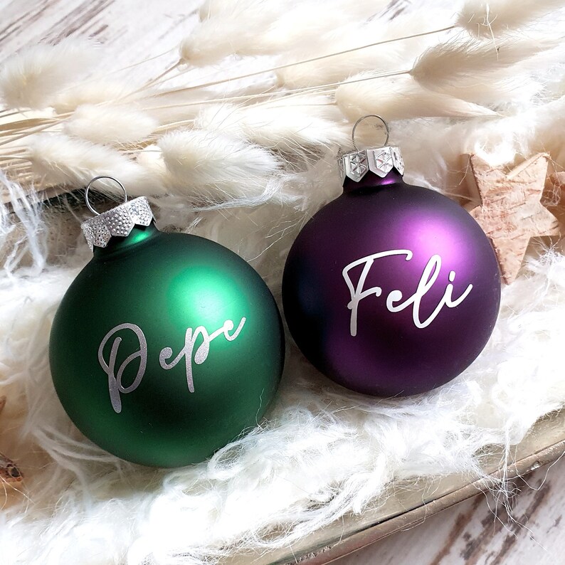 May include: Two Christmas ornaments, one green with the name "Pepe" written in white and one purple with the name "Feli" written in white.