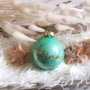 May include: A green glass Christmas ornament with a gold top and the name "Nina" written in gold lettering.