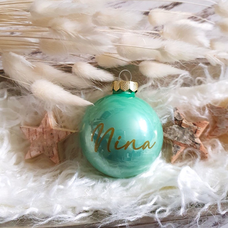 May include: A green glass Christmas ornament with a gold top and the name "Nina" written in gold lettering.