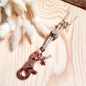 May include: A beige and rose gold key chain with a star charm and a beige rope with a beige bead. The key chain has a rose gold lobster clasp.