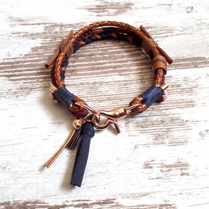 May include: A braided leather dog collar with a brown, blue, and burgundy color scheme. The collar has a gold-toned metal clasp and a tassel charm.