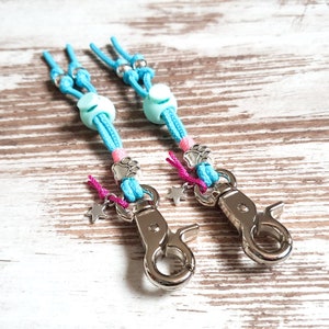 May include: Two turquoise blue and pink keychains with silver clips. Each keychain has a turquoise blue bead, a pink paw print charm, and a silver star charm.