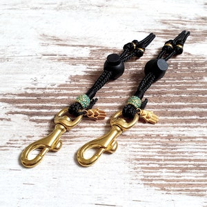 May include: Two black braided lanyards with gold-colored clasps and decorative beads. Each lanyard has a small gold hand charm and turquoise-colored beads. The clasps are attached to the braided cord.