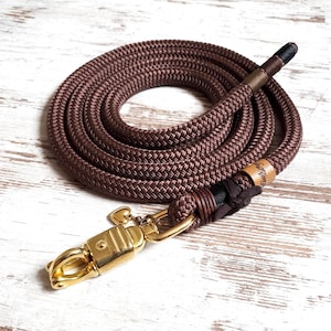 Op de afbeelding: Een bruine touwhalster voor honden met een gouden metalen sluiting en een klein hartvormig sieraad. De halster is opgerold en heeft een accent van bruin leer en zwart garen.