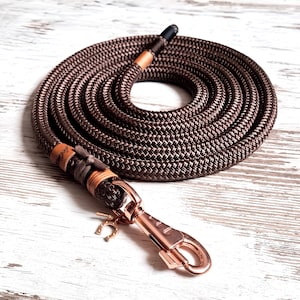 Op de afbeelding: Een bruine touwhalster voor honden met een roségouden haak en een hoefijzerbedel. De halster is opgerold op een wit houten oppervlak.
