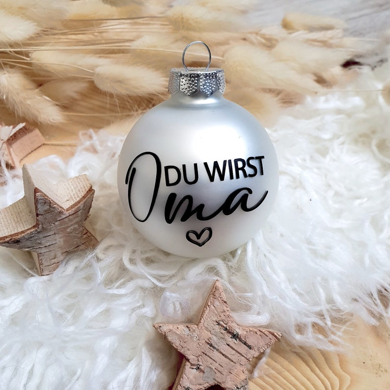 May include: A silver Christmas ornament with the text "DU WIRST Oma" in black lettering and a small heart.