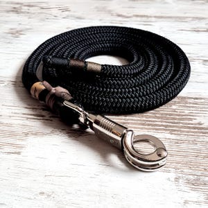 Op de afbeelding: Zwarte touwriem opgerold op een wit houten oppervlak. De riem heeft een zilveren metalen sluiting en decoratieve bruine en beige accenten. Het touw is dik en lijkt duurzaam, geschikt voor huisdieren.