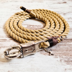 Op de afbeelding: Een opgerolde beige touwen hondenriem met een zilveren metalen sluiting. De riem heeft bruine lederen accenten en een kleine hondenbedel. Het touw is dik en lijkt duurzaam.