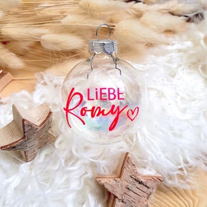 May include: A clear glass Christmas ornament with a red heart and the text "LIEBE Romy" in red lettering.