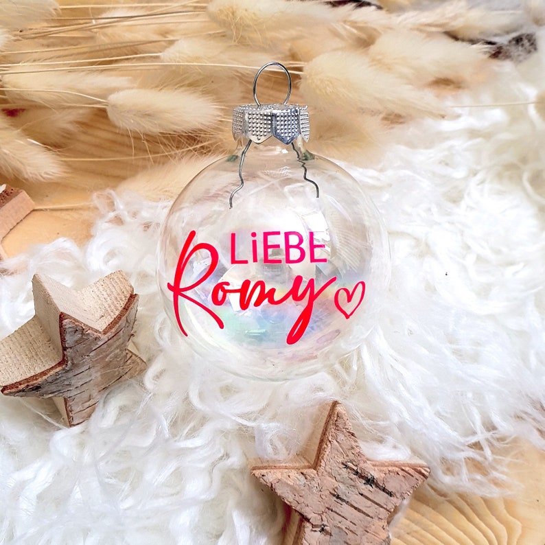 May include: A clear glass Christmas ornament with a red heart and the text "LIEBE Romy" in red lettering.