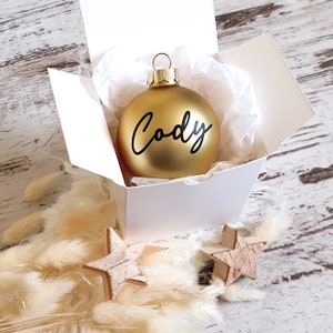 May include: A gold Christmas ornament with the name "Cody" written in black script. The ornament is in a white box with tissue paper.