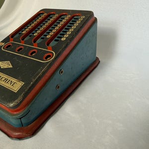 May include: A vintage Wolverine metal adding machine toy with a blue body, red trim, and a black top panel. The device features vertical slots with numbered dials from zero to nine, four circular openings, and the text Wolverine Made in USA.