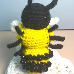May include: A handmade crochet bee with a black head and antennae, a yellow and black striped body, and black bead accents. The bee has black legs and white wings. The craft item is sitting on a white, lacy surface.