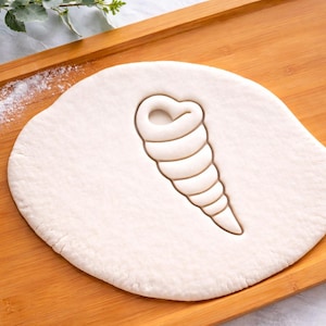 May include: A seashell-shaped cookie cutter pressed into a round of dough, resting on a wooden cutting board. The dough is light in color, and the cutter has created a detailed seashell impression. Flour is lightly sprinkled on the board.