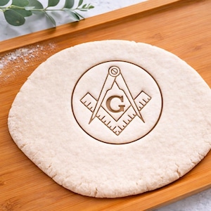 May include: A round of dough with a Masonic symbol pressed into it, featuring a compass, square, and the letter "G". The dough sits on a wooden cutting board, with flour and greenery in the background.
