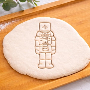 May include: A light-colored dough with a nutcracker imprint on a wooden tray. The nutcracker design is detailed, showing the figure's uniform and facial features. Flour is sprinkled on the tray.