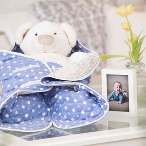 May include: A white teddy bear wrapped in a blue and white polka dot blanket. The blanket has a white border and a white ribbon tied around it. A small white picture frame with a photo of a baby boy is on the table next to the teddy bear.