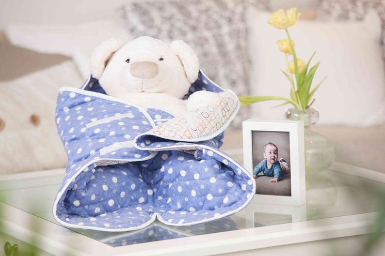 May include: A white teddy bear wrapped in a blue and white polka dot blanket. The blanket has a white border and a white ribbon tied around it. A small white picture frame with a photo of a baby boy is on the table next to the teddy bear.