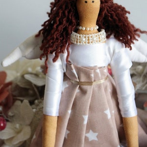Puede incluir: Una mu&ntilde;eca hecha a mano con cabello rizado marr&oacute;n, un vestido blanco con un collar de perlas y una falda marr&oacute;n con estrellas blancas. La mu&ntilde;eca tiene un ala blanca en un lado.