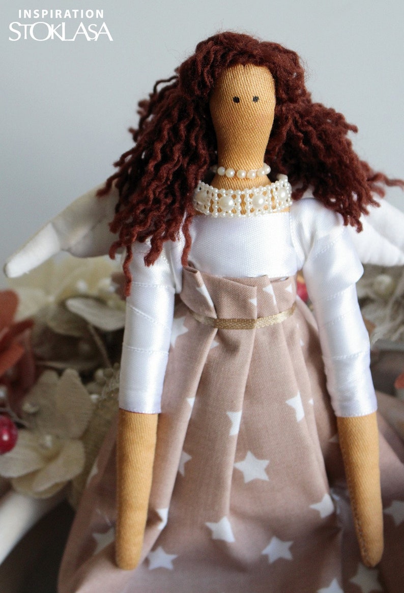 Puede incluir: Una mu&ntilde;eca hecha a mano con cabello rizado marr&oacute;n, un vestido blanco con un collar de perlas y una falda marr&oacute;n con estrellas blancas. La mu&ntilde;eca tiene un ala blanca en un lado.