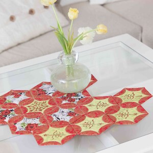 May include: A red and green patchwork table runner with a floral design. The runner is on a glass-topped coffee table with a vase of yellow tulips.