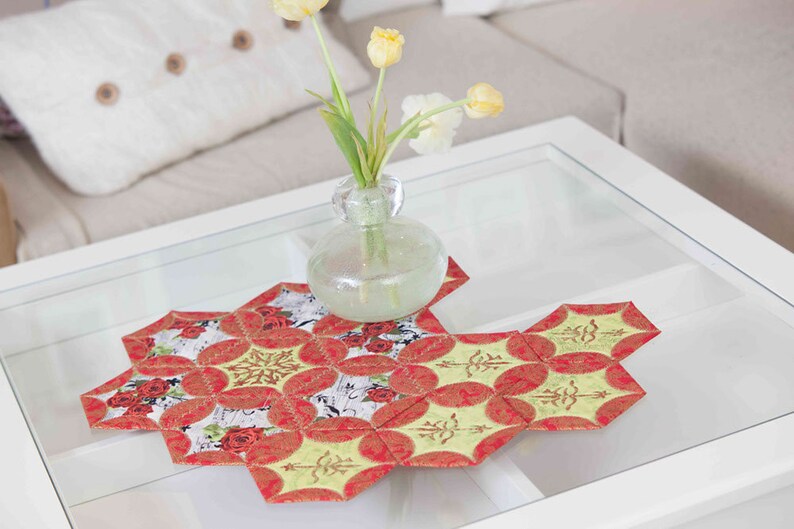 May include: A red and green patchwork table runner with a floral design. The runner is on a glass-topped coffee table with a vase of yellow tulips.